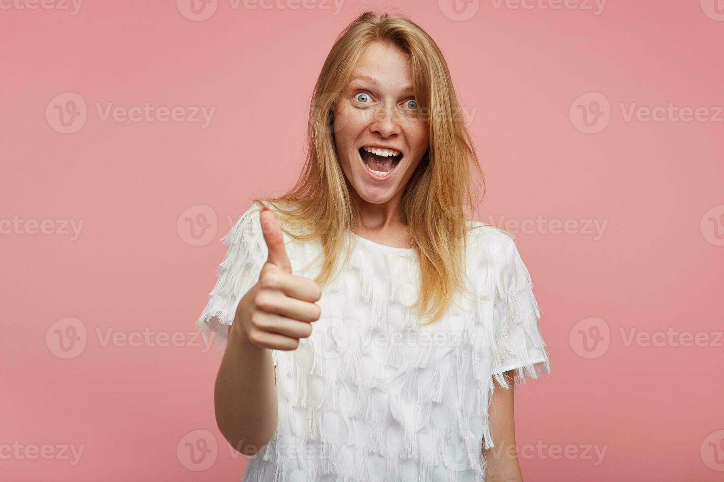 Overjoyed young lovely female with foxy hair showing hand with raised thumb and looking excitedly at camera with opened mouth, standing against pink background photo