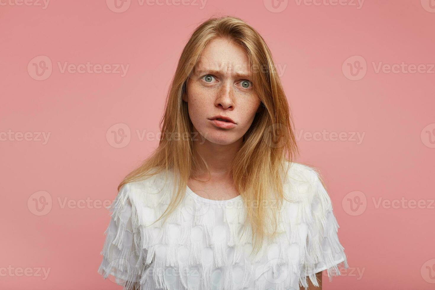 Puzzled young lovely redhead lady with natural makeup frowning her eyebrows while looking amazedly at camera, dressed in elegant clothes while standing over pink background photo