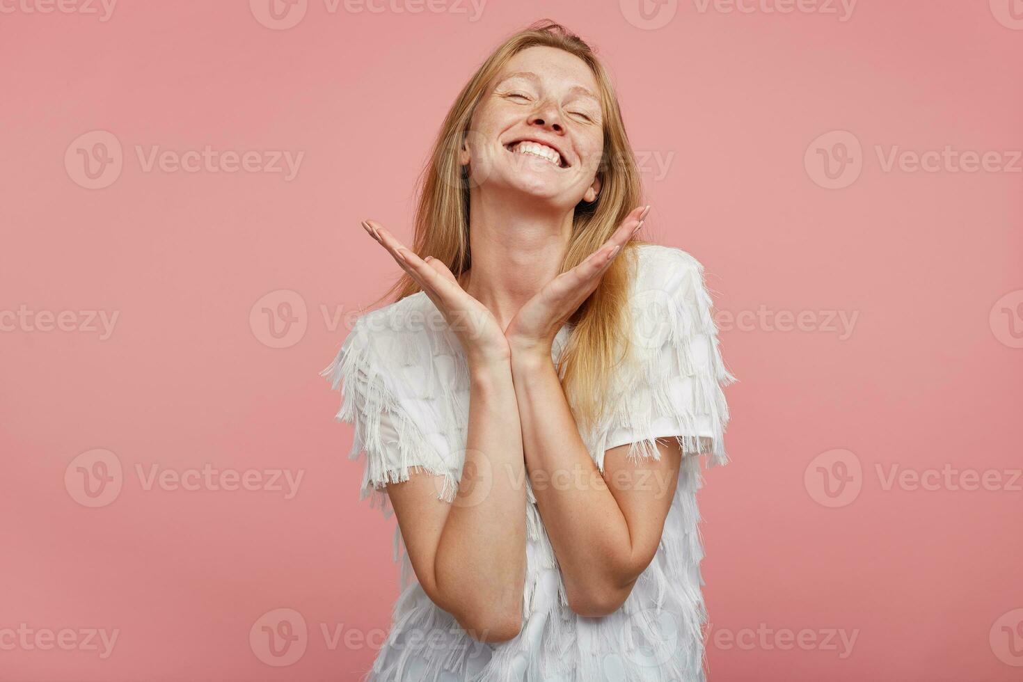 Pleasant looking young cheerful woman with foxy hair keeping raised hands under her head and smiling widely with closed eyes, wearing white festive t-shirt while posing over pink background photo