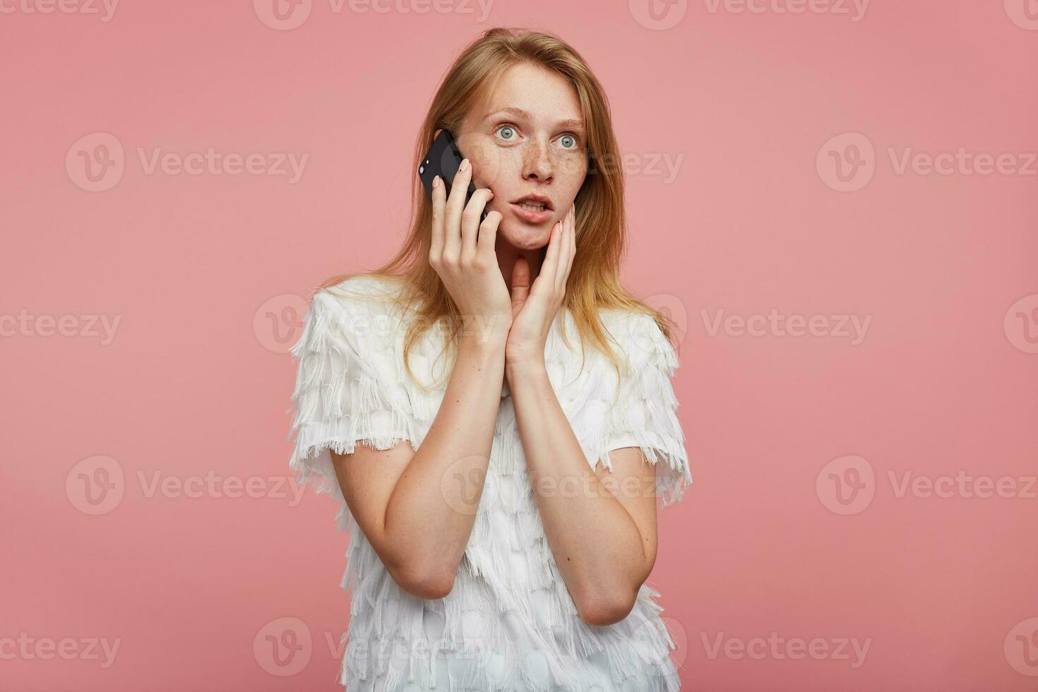 Studio photo of open-eyed pretty young redhead lady holding palm on her face while posing over pink background, looking surprisedly ahead while having phone conversation