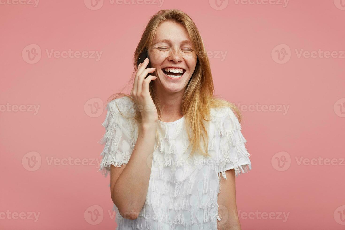 Good looking young lovely redhead lady with natural makeup keeping her eyes closed while laughing happily, having phone talk while standing over pink background photo