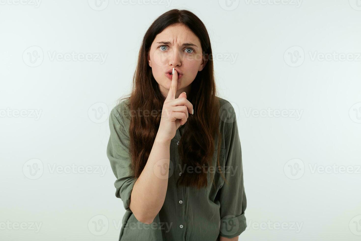 Severe young brunette woman with casual hairstyle frowning her eyebrows while looking seriously at camera and raising hand in hush gesture, isolated over white background photo