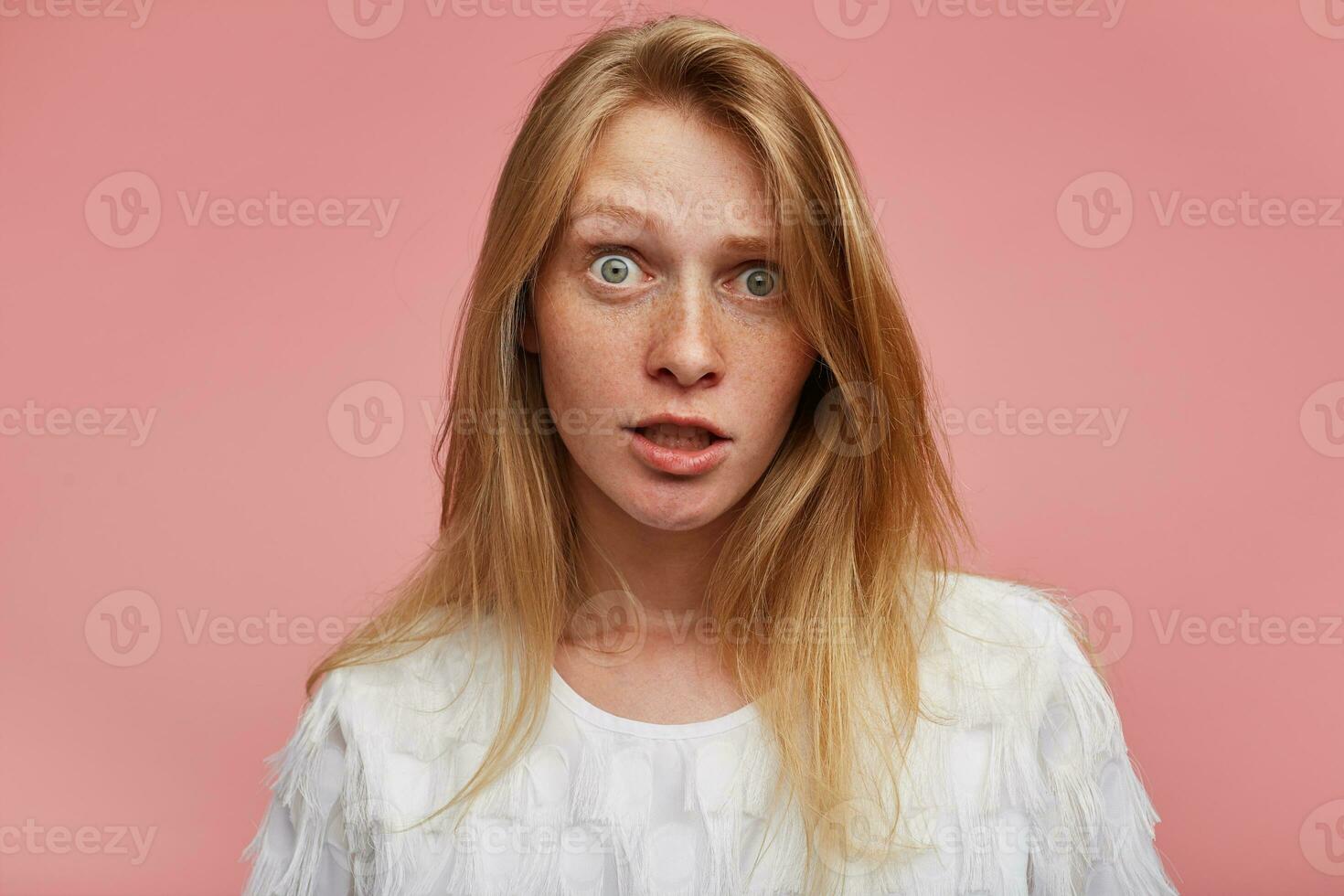 Close-up of confused young redhead lady with casual hairstyle looking at camera with wide eyes opened while standing over pink background in white festive t-shirt photo