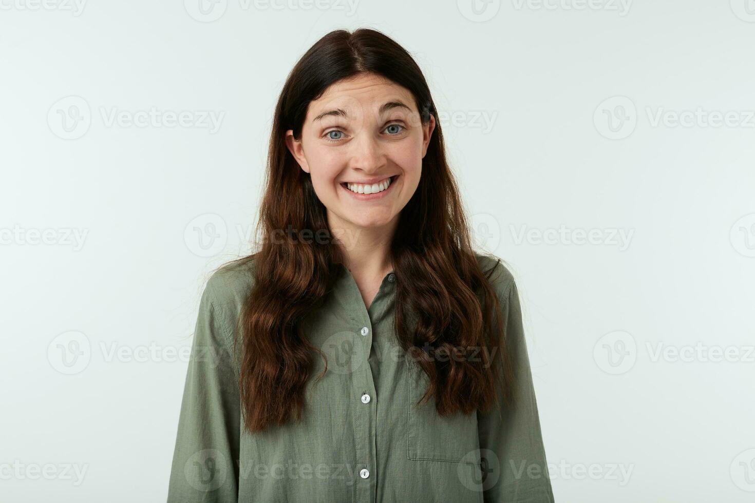 Studio photo of young happy brunette female with long wavy hair smiling widely while looking joyfully at camera, isolated over white background in casual shirt