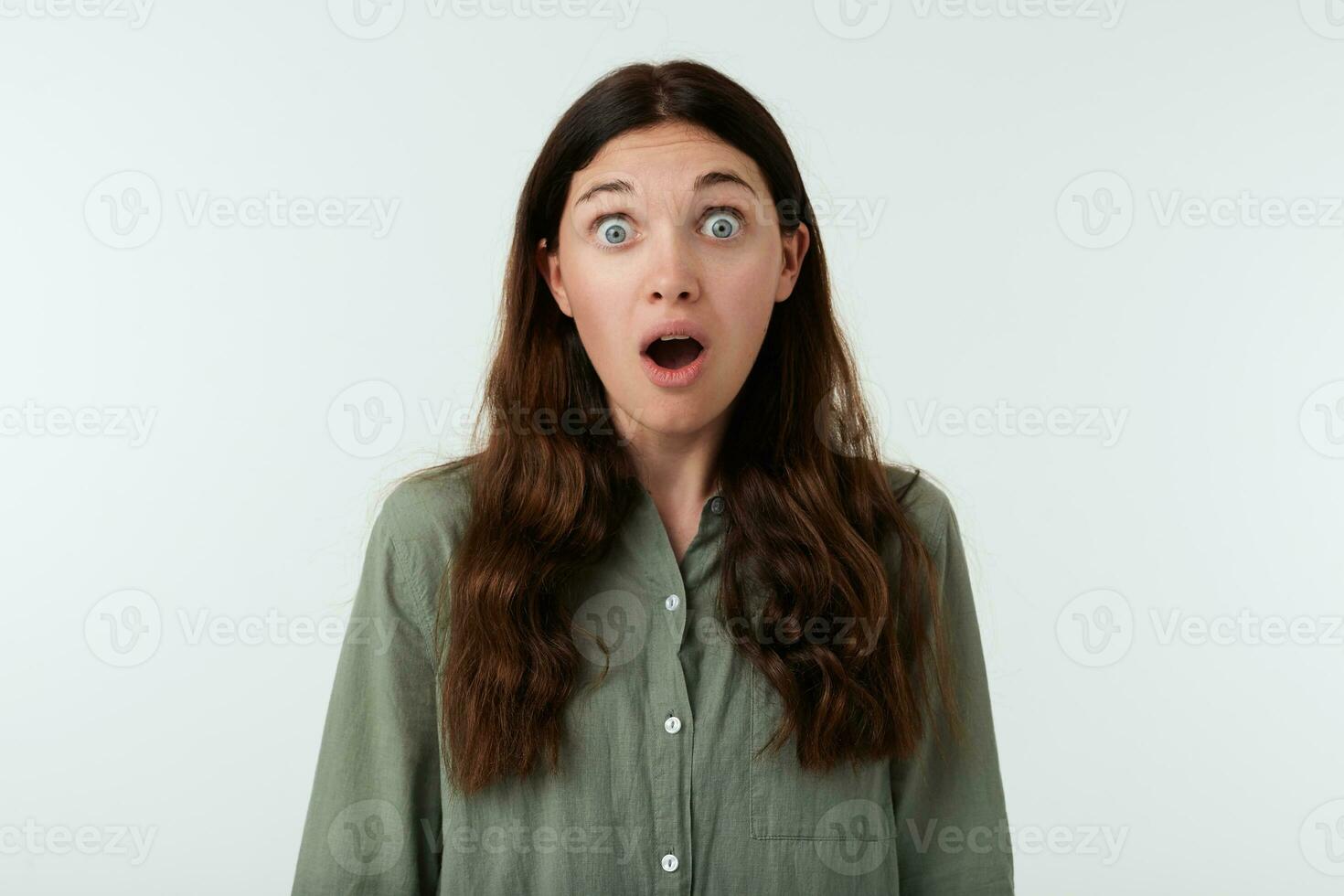 Frightened young pretty brown haired woman with natural makeup looking amazedly at camera with wide eyes and mouth opened, standing over white background photo