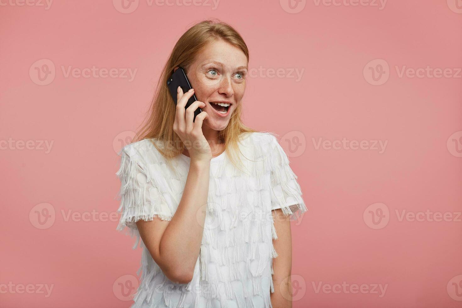 Agitated young attractive female with natural makeup looking joyfully ahead with excited face while making call to her friend, standing against pink background photo