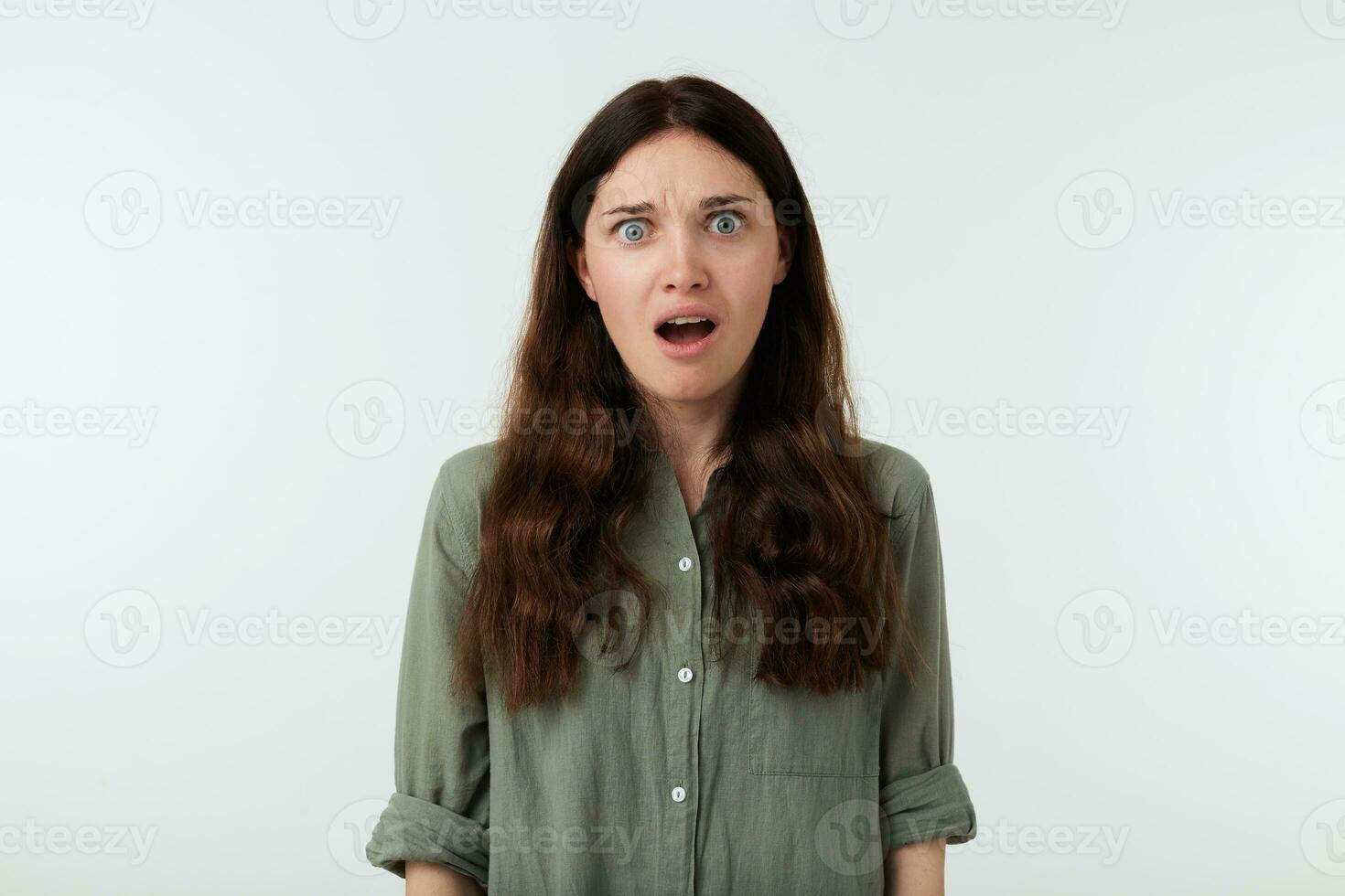 Bemused young long haired brunette lady with wavy hairstyle grimacing her face while looking amazedly at camera with opened mouth, isolated over white background photo