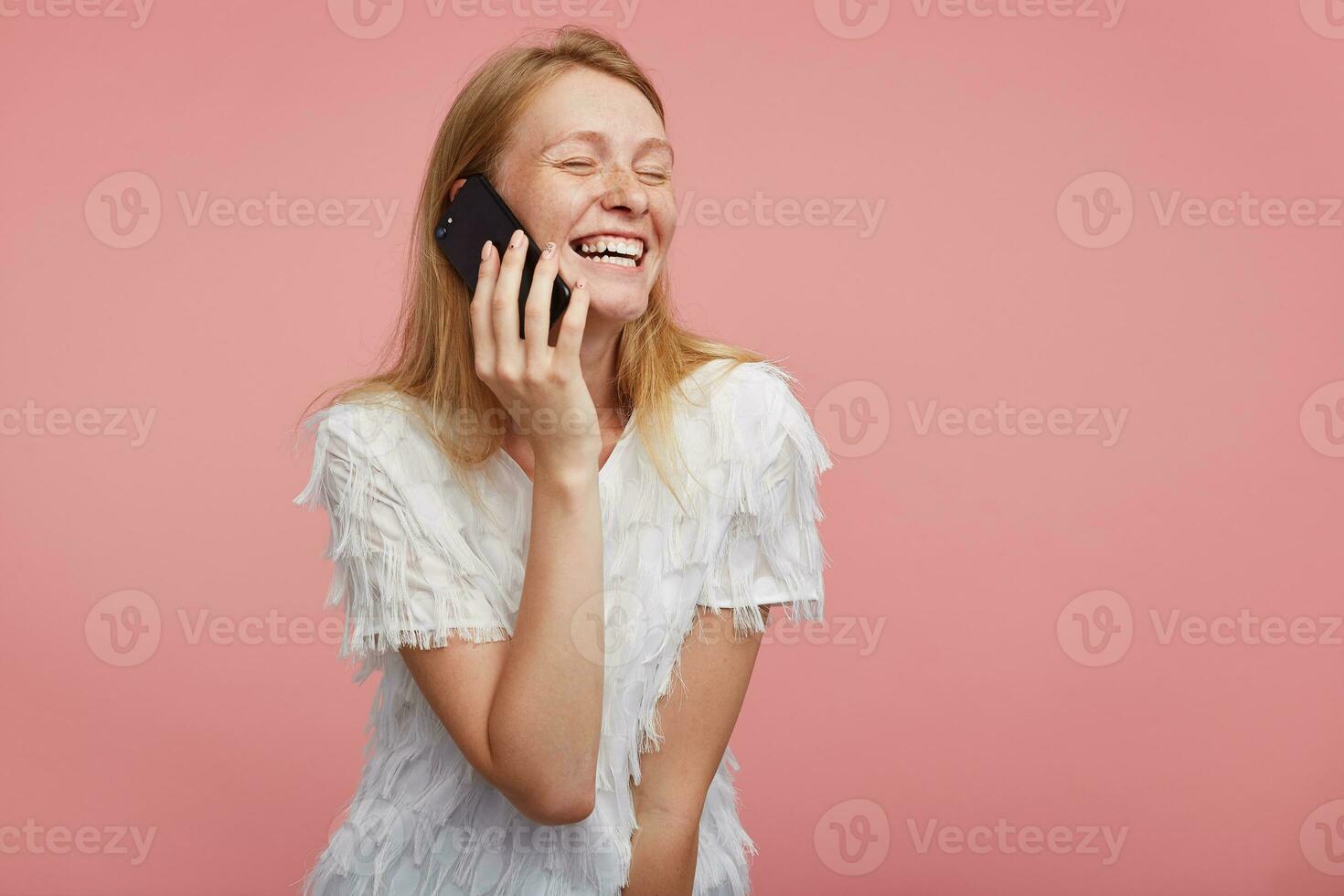 Studio photo of joyful young pretty redhead lady smiling widely with closed eyes while having pleasant talk on phone, wearing festive t-shirt while posing over pink background