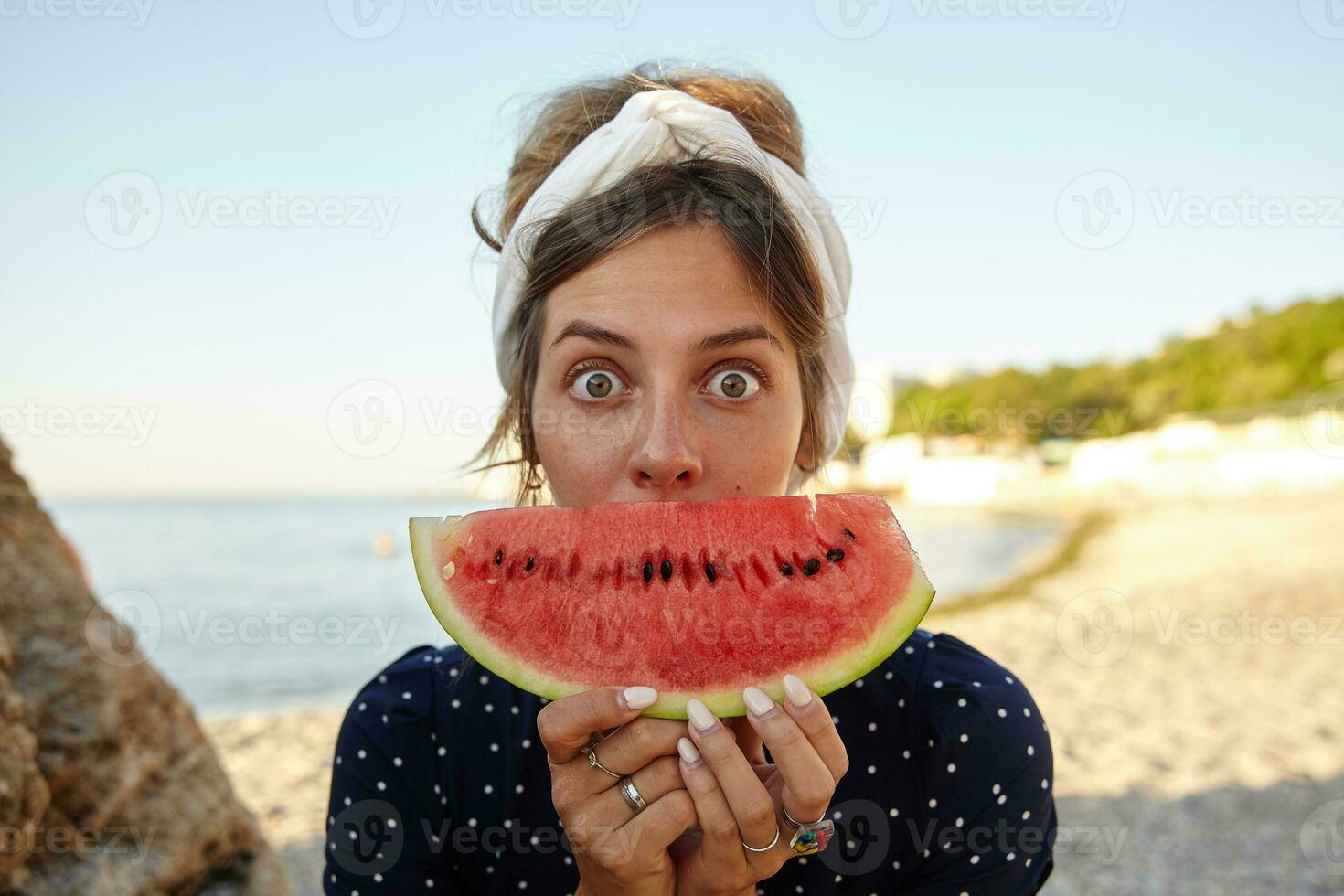 Young dark haired pretty woman in headband making fun over seaside view with slice of watermelon in her hands, looking at camera with wide eyes opened and raising eyebrows photo