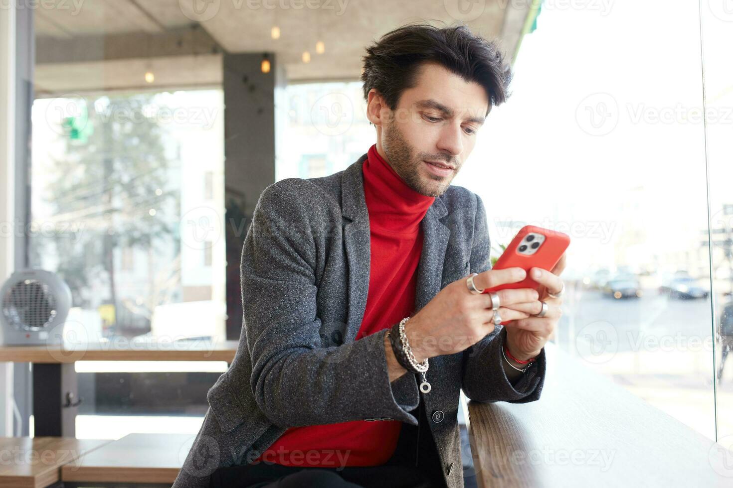 Elegant young pretty dark haired bearded man keeping mobile phone in his hands and looking attentinvely on screen while being isolated over urban background photo