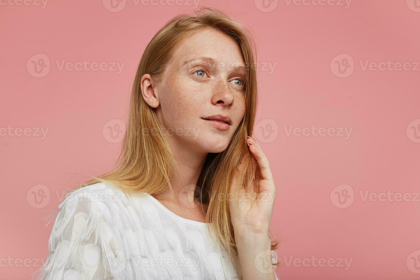 Studio shot of young beautiful redhead lady with green-gray eyes raising hand to her face while looking dreamily aside, dressed in white t-shirt while posing over pink background photo