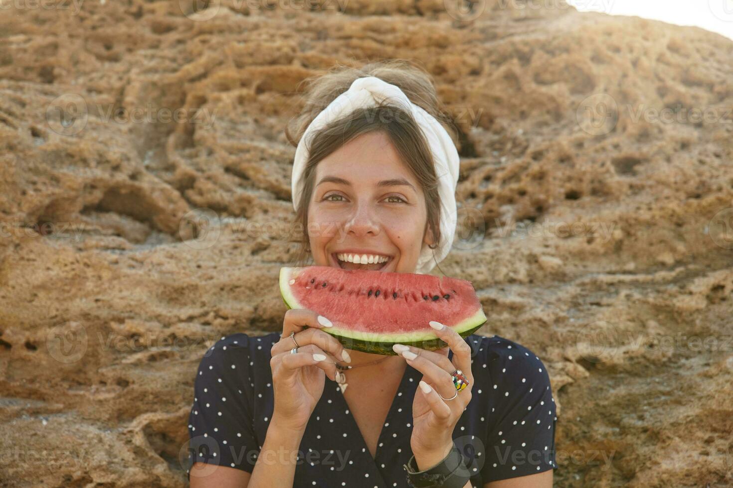 Happy beautiful young dark haired female in headband with casual hairstyle posing over big yellow stone on sunny warm day, eating watermelon and looking cheerfully to camera photo