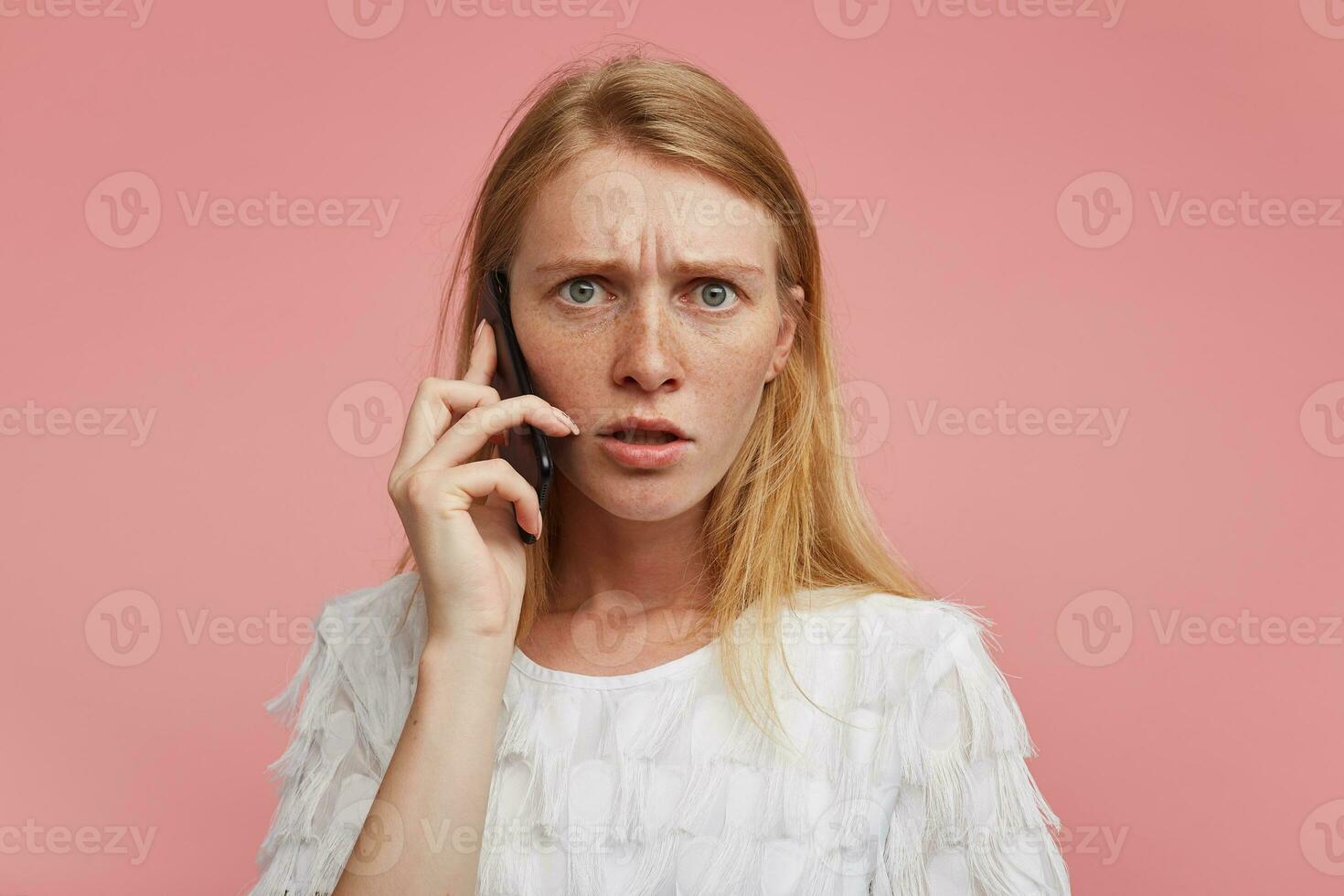 Severe young pretty lady with foxy hair frowning her eyebrows while having phone talk and looking at camera with serious face, wearing white elegant t-shirt while posing over pink background photo