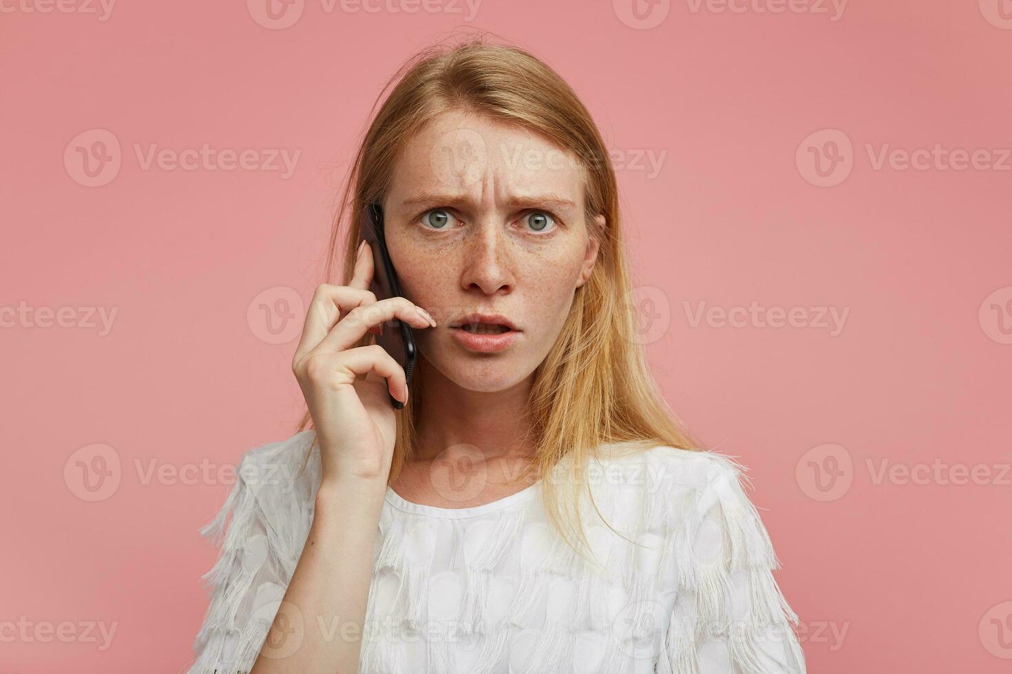 grave joven bonito dama con astuto pelo ceñudo su Cejas mientras teniendo teléfono hablar y mirando a cámara con grave rostro, vistiendo blanco elegante camiseta mientras posando terminado rosado antecedentes foto