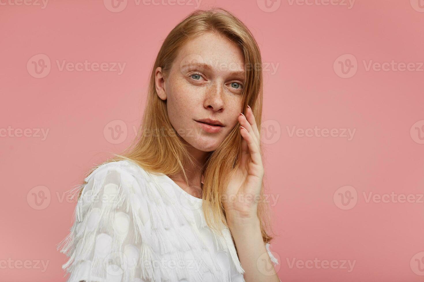 Closeup of young pensive attractive redhead woman with natural makeup touching soft her face with raised hand and looking at camera with calm face, isolated over pink background photo