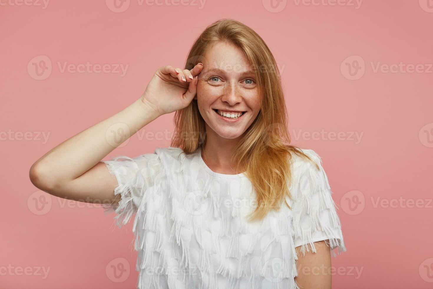 Studio shot of cheerful beautiful young female with foxy hair touching gently her face with raised hand and looking positively at camera with broad smile, isolated over pink background photo