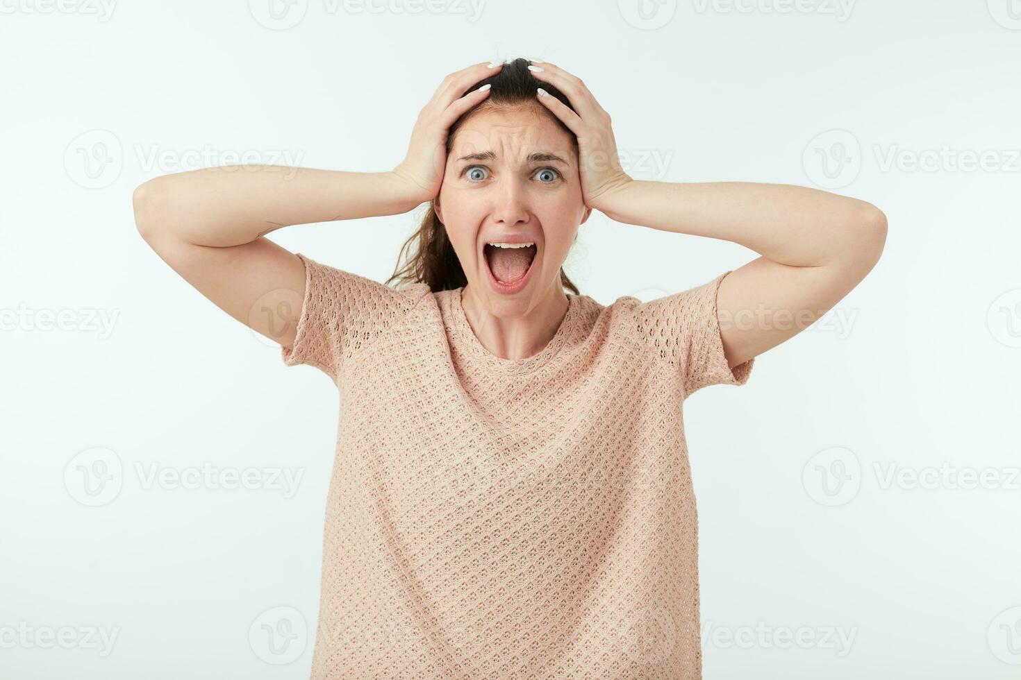 Stressed young dark haired woman with ponytail hairstyle clutching her head with raised hands while screaming with wide mouth opened, isolated over white background photo