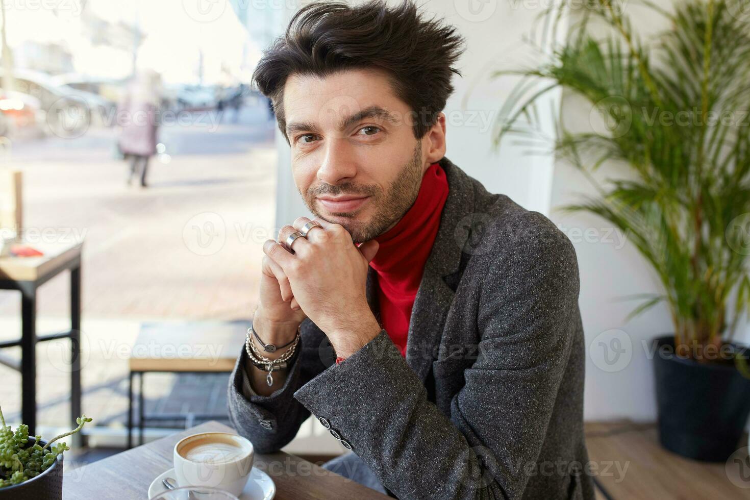 Portrait of beautiful young bearded brunette man looking positively at camera with charming smile and leaning his head on raised hands while sitting in city cafe photo