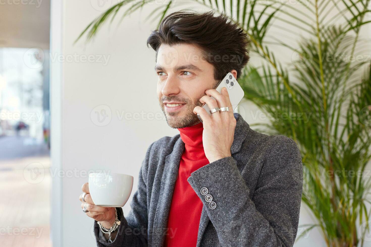 Good looking young glad unshaved brunette male keeping cup of coffee in raised hand and smiling pleasantly, making call while posing over cafe interior photo