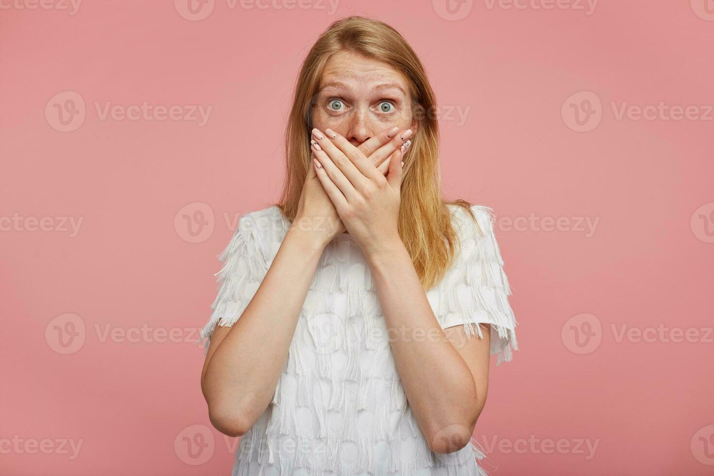 Open-eyed young pretty lady with foxy hair raising surprisedly eyebrows while looking amazedly at camera, covering mouth with raised hands while posing over pink background photo