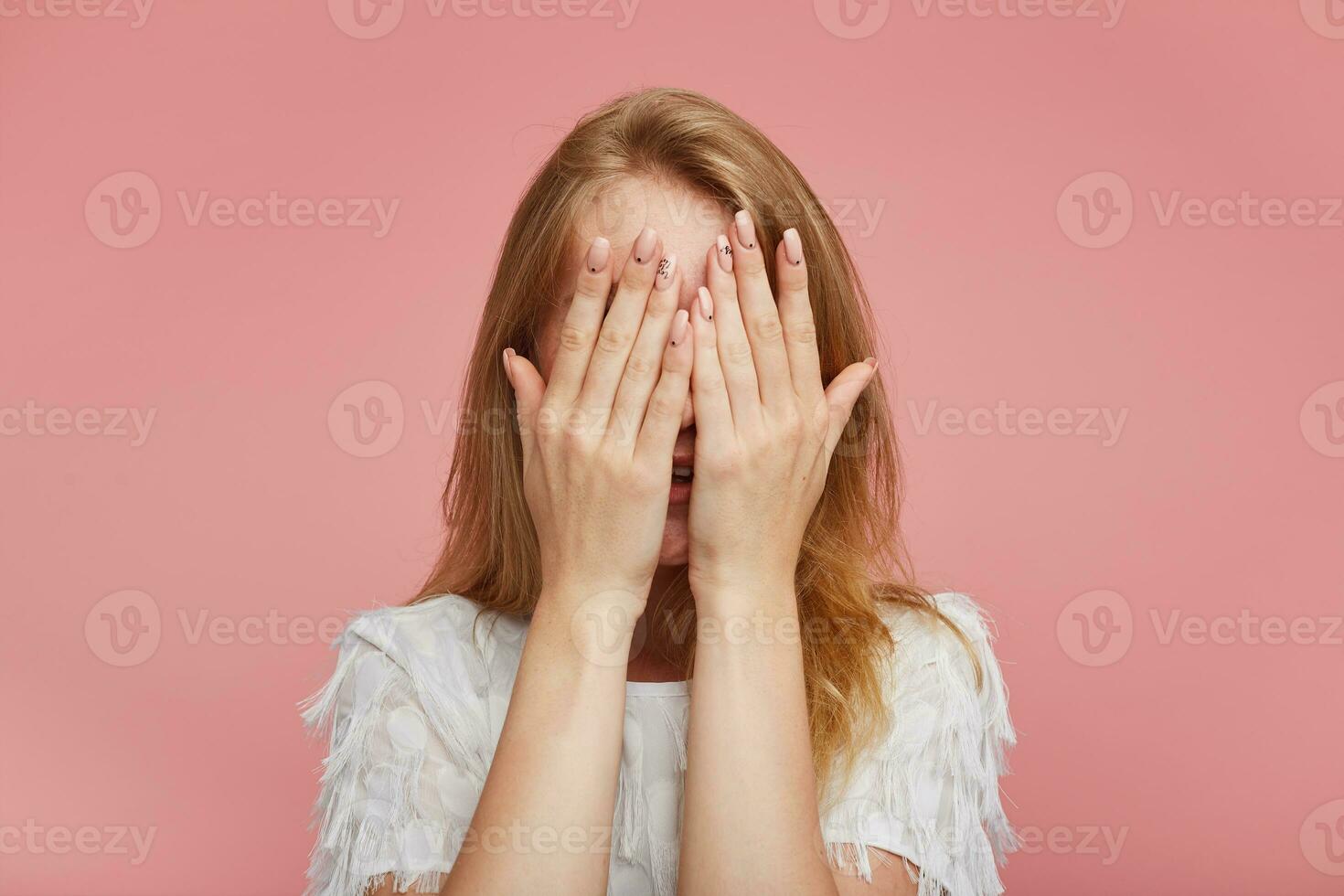 Closeup of young redhead woman with casual hairstyle keeping raised hands with nude manicure in front of her face, wearing white elegant t-shirt while posing over pink background photo