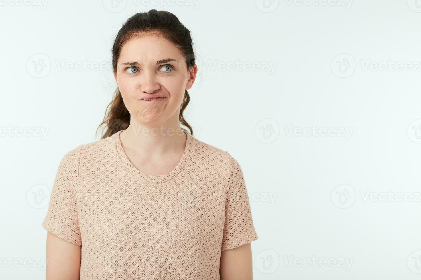 Horizontal shot of young green-eyed pretty brunette female grimacing her face while looking confusedly aside, standing over white background with hands down photo