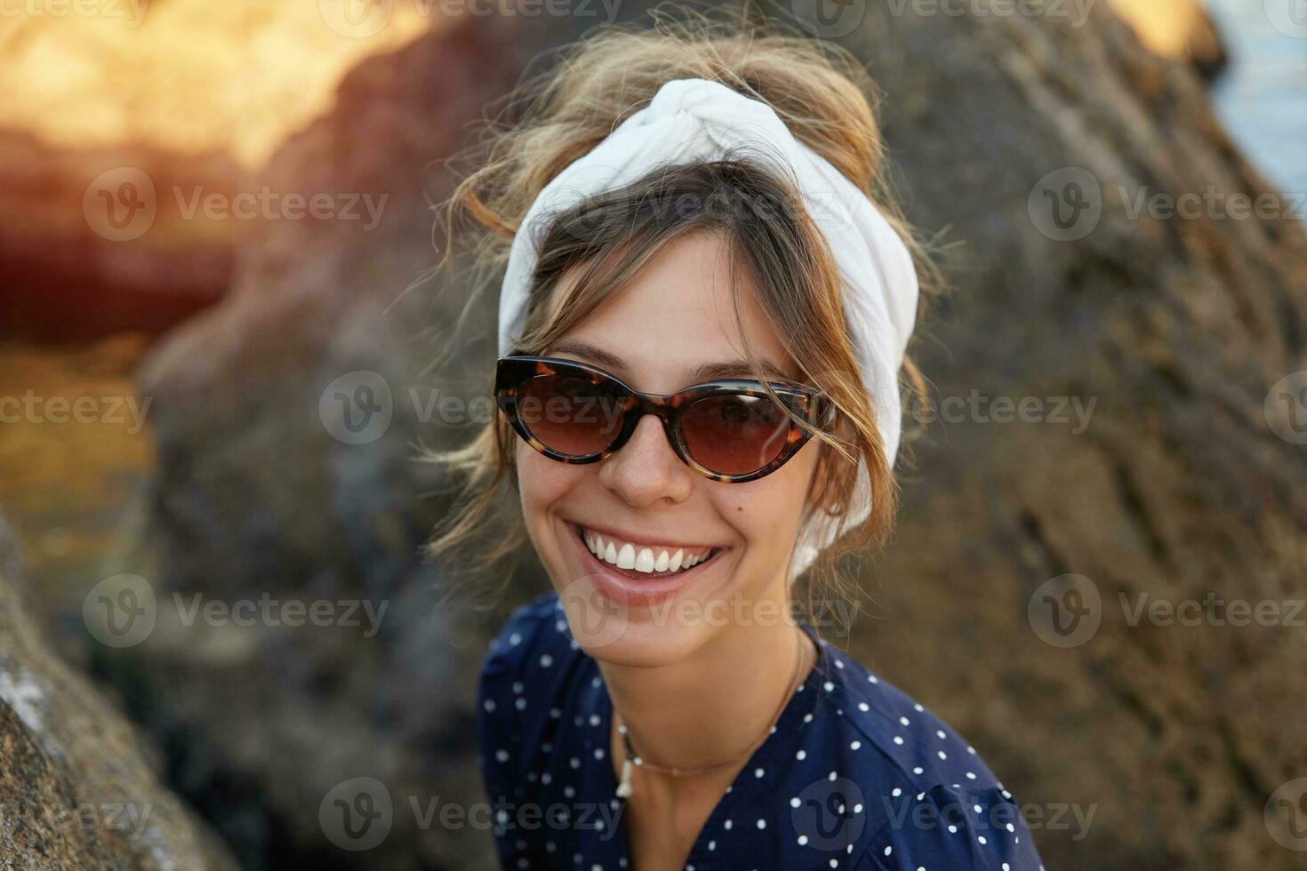 Outdoor shot of young cheerful female in vintage eyewear wearing romantic dress, looking at camera with happy wide smile, posing over seaside on summer day photo