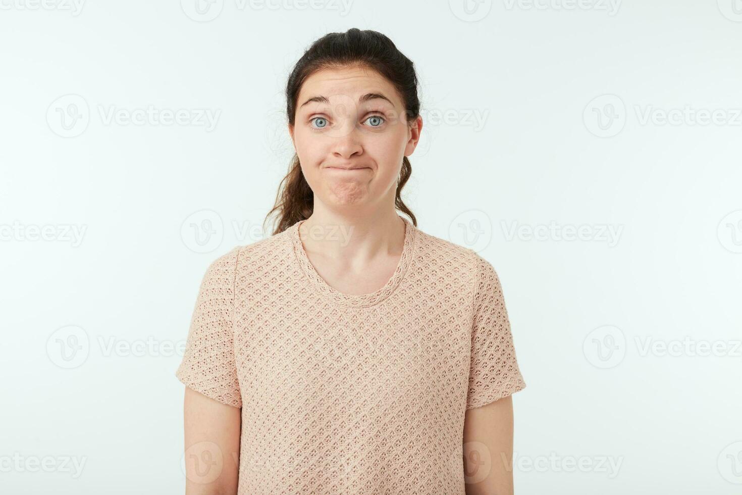 Indoor photo of young dark haired woman with natural makeup making bewildered face while looking at camera while standing against white background in casual wear
