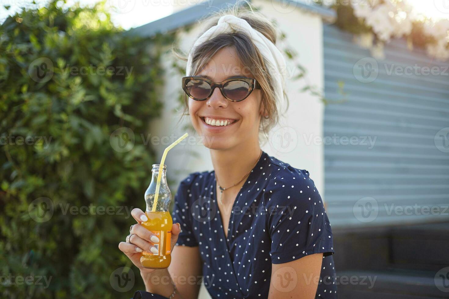 Portrait of pretty young lady in trendy eyewear wearing romantic dress while sitting outdoor with lemonade in raised hand, looking at camera with charming smile photo