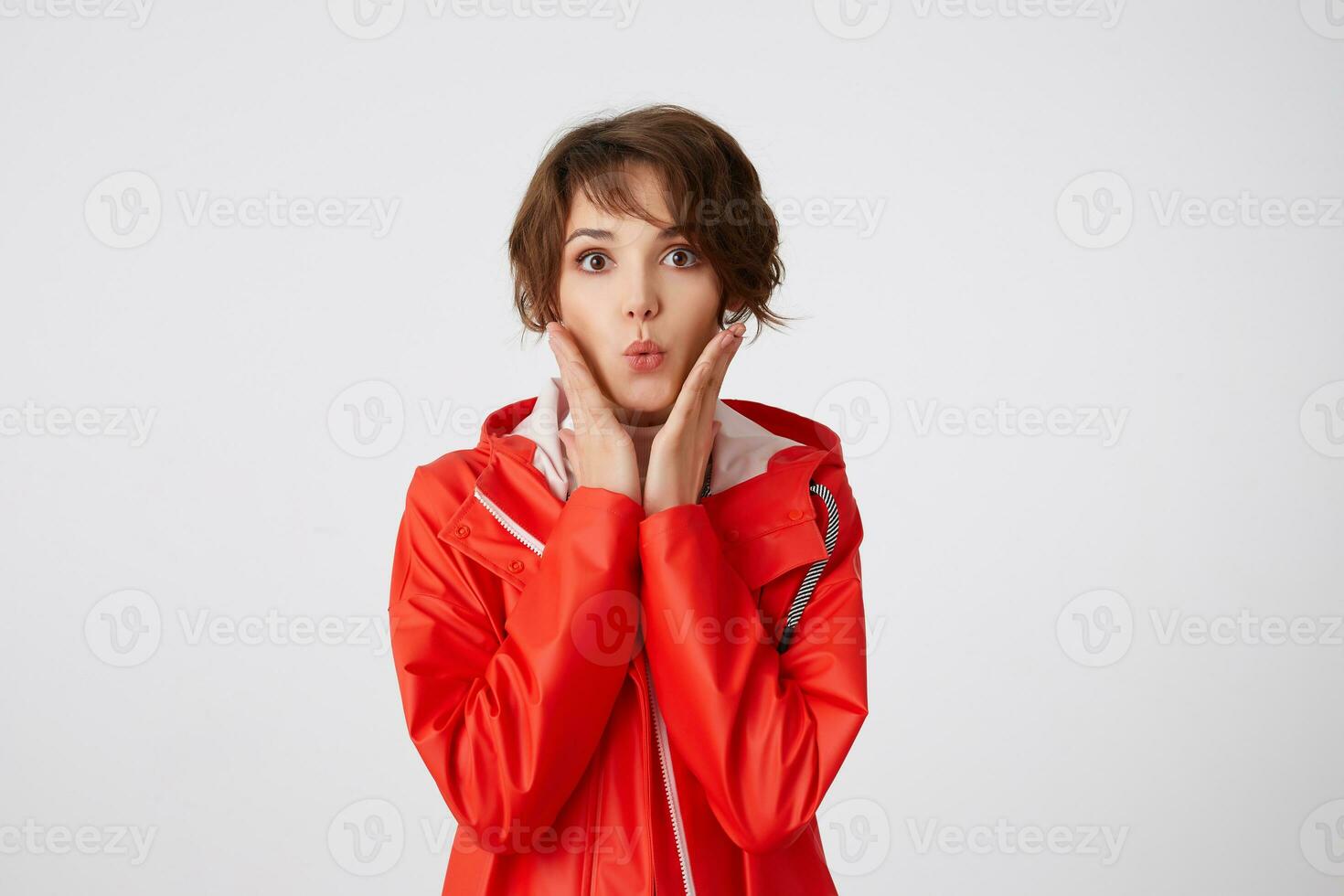 Photo of young cute short haired girl dressed in white golf and red rain coat, looking at the camera with amazed expression, send kiss, palms on cheeks, standing over white background.