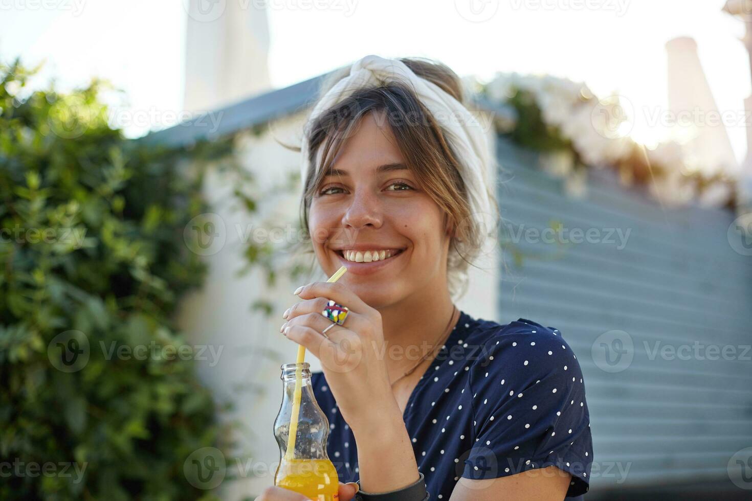 hermosa joven mujer en blanco venda vistiendo romántico vestir mientras posando al aire libre en calentar soleado día, mirando a cámara alegremente y sonriente ampliamente, Bebiendo naranja jugo con Paja foto