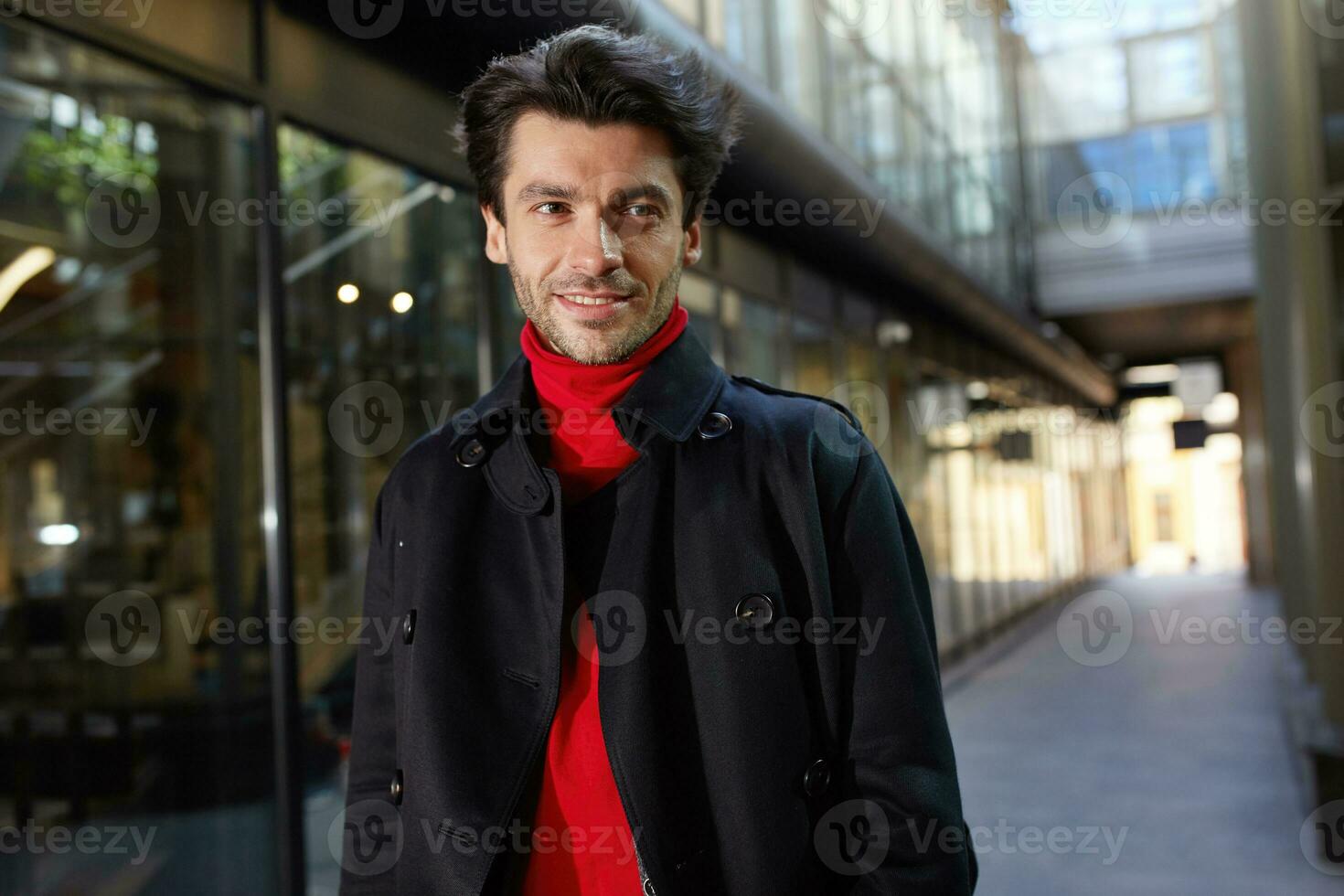 retrato de joven bonito marrón peludo barbado chico sonriente alegremente mientras caminando al aire libre en soleado día, vestido en formal ropa mientras posando terminado ciudad antecedentes foto