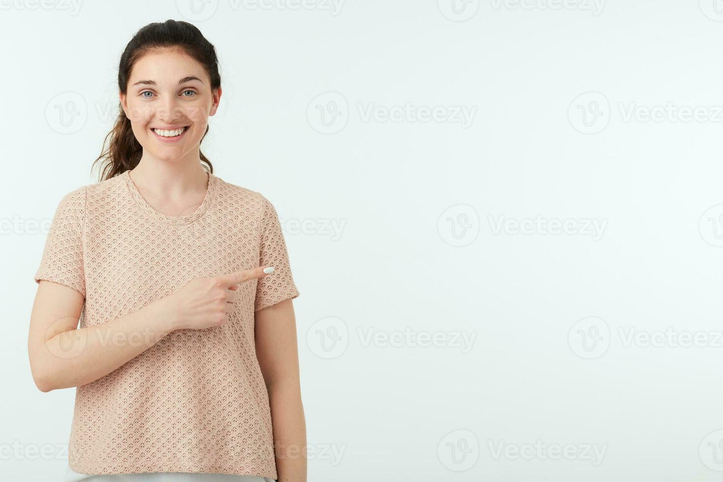 Pleased young beautiful dark haired lady with ponytail hairstyle keeping her hand raised while pointing aside with forefinger and smiling gladly at camera, isolated over white background photo