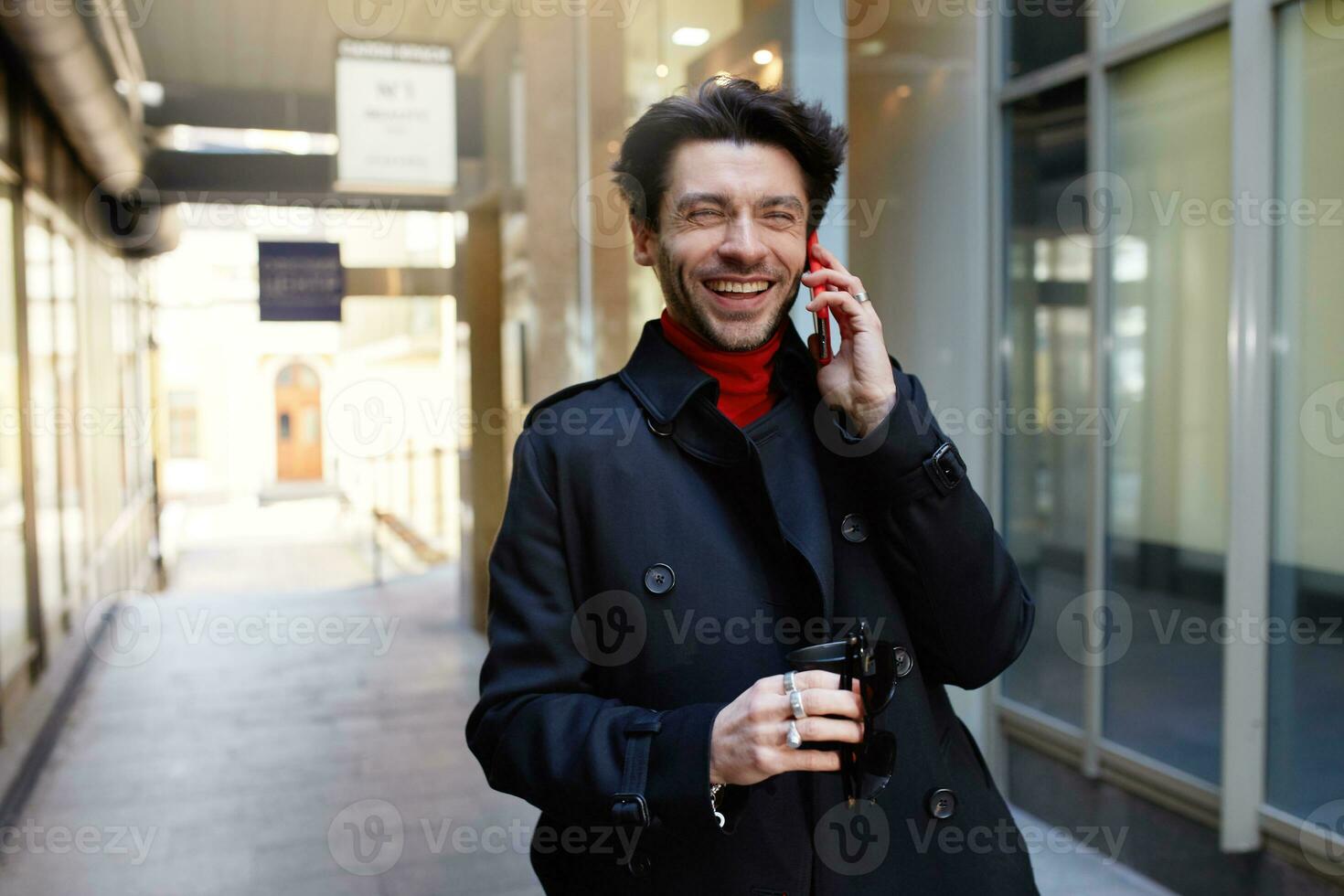 Joyful young attractive brown haired male with trendy haircut laughing happily while having phone conversation with friend, isolated over urban background photo