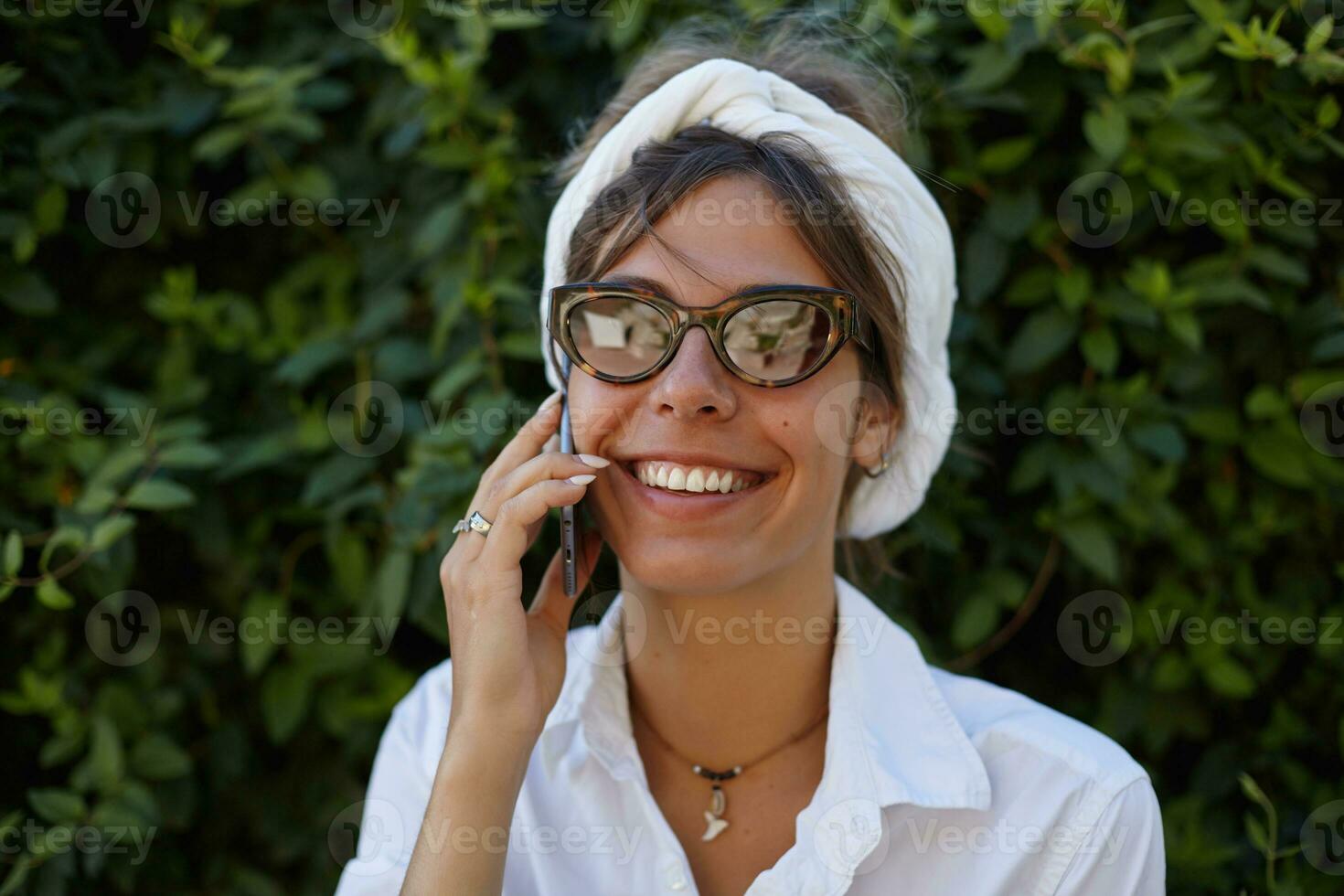 Joyful attractive young lady in vintage eyewear and white headband keeping smartphone in her hand and making call, smiling happily to camera while posing over green garden photo