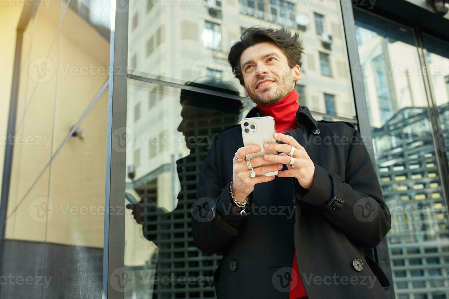 Horizontal shot of cheerful young handsome dark haired unshaved man looking gladly ahead while holding smartphone in raised hands, isolated over city background photo