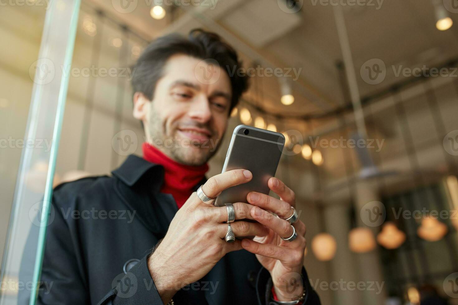 Close-up of raised man's hands with rings keeping mobile phone while posing over city cafe background, texting message to friends and smiling slightly while looking positively on screen photo