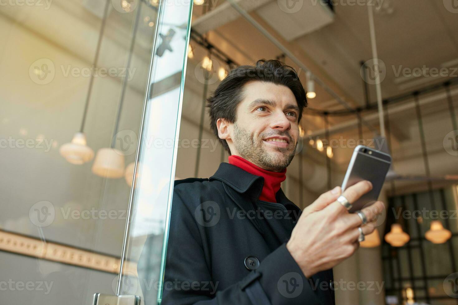 Pleasant looking young attractive dark haired man smiling gladly while keeping smartphone in raised hand, posing over cafe interior in elegant clothes photo