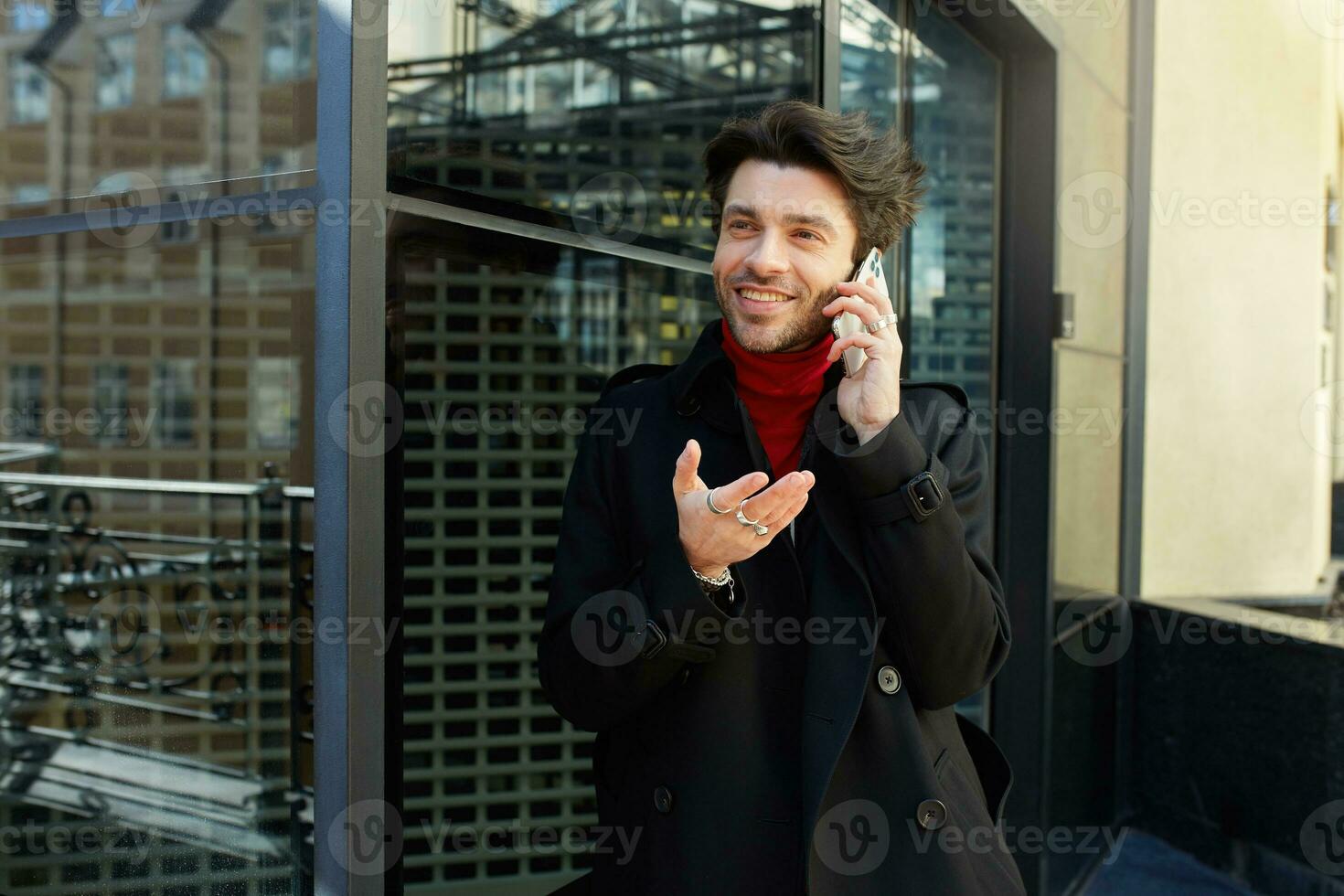 Glad young pretty brown haired man with trendy haircut raising emotionally palm while havingphone conversation and smiling happily, posing outdoor on sunny day photo