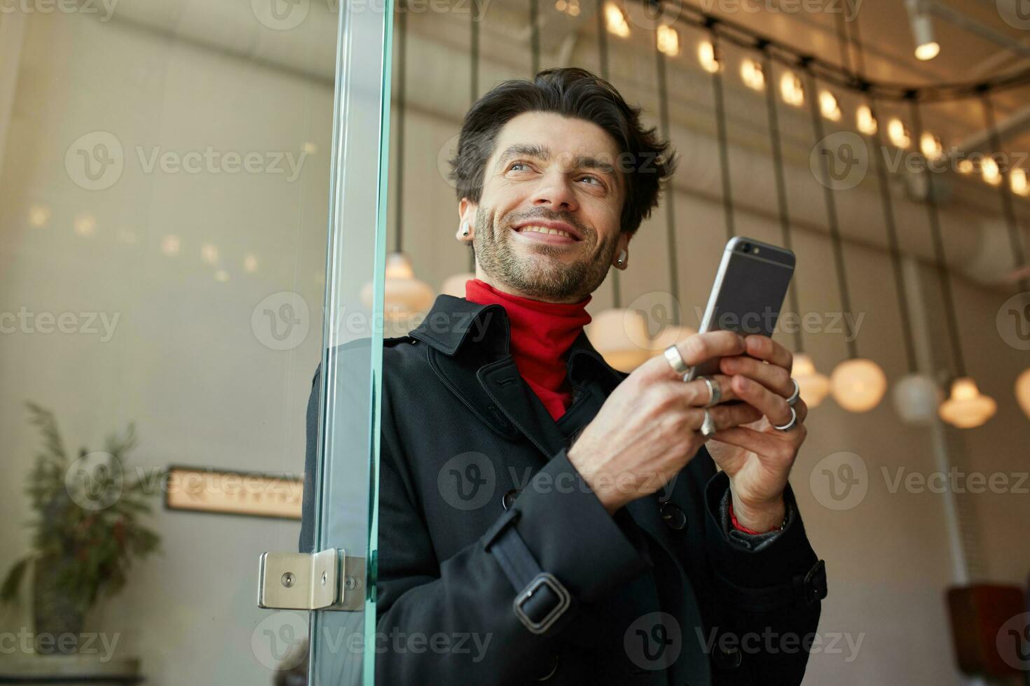 Pleased young handsome brown haired with earpieces keeping smartphone and looking cheerfully aside with wide smile, standing over city cafe background photo
