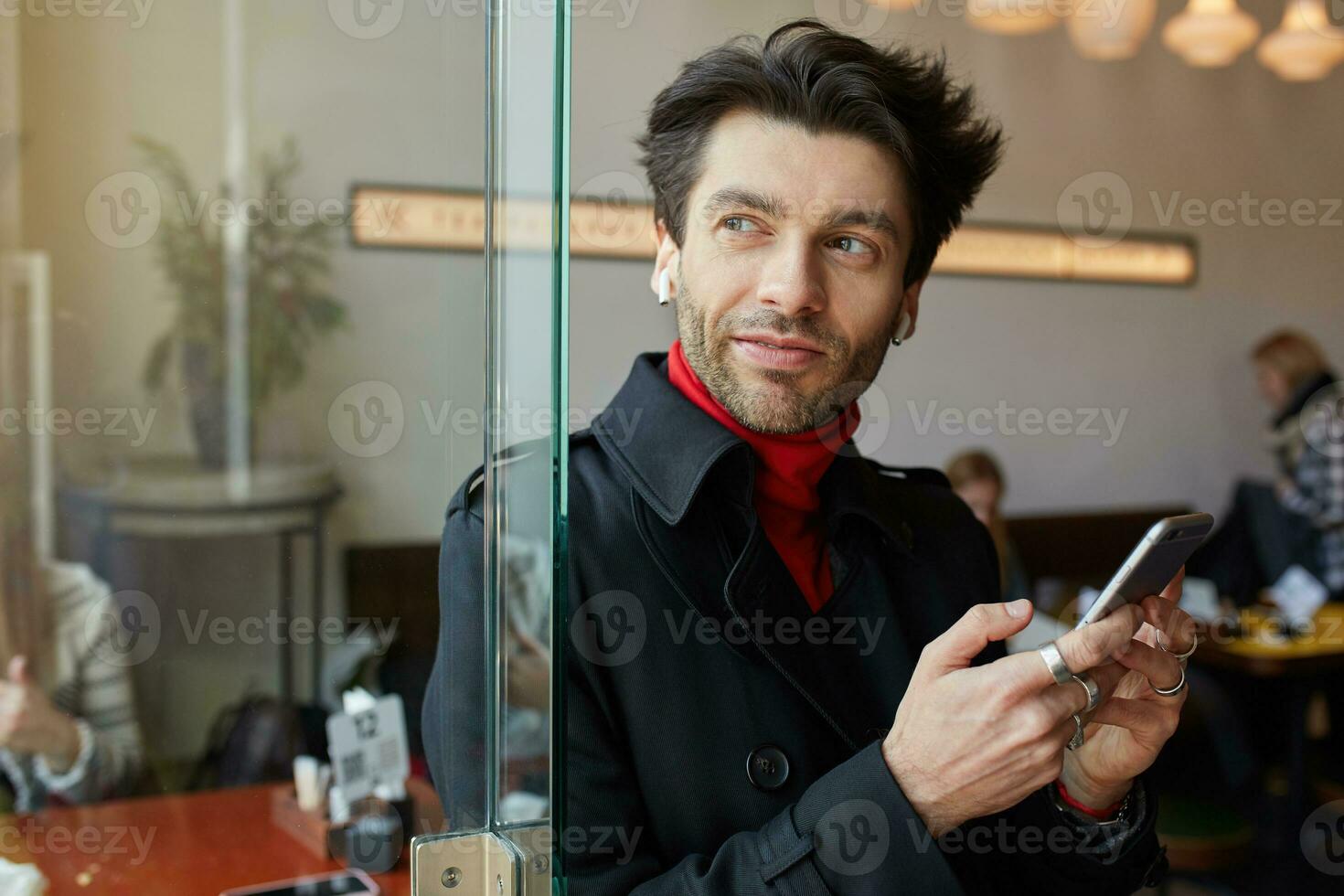 positivo joven hermosa marrón peludo barbado chico acuerdo móvil teléfono en elevado manos y mirando pensativamente en ventana, posando terminado café casa antecedentes foto