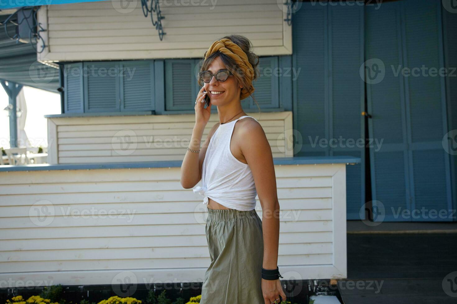 Attractive young brunette female with casual hairstyle wearing white top and olive skirt walking outdoor on summer warm day, holding mobile phone in raised hand and making call to friend photo