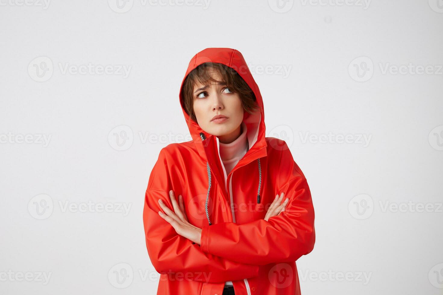 Young unhappy cute short haired lady dressed in red rain coat, with a hood on his head, looking away with sad expression, standing over white background. photo