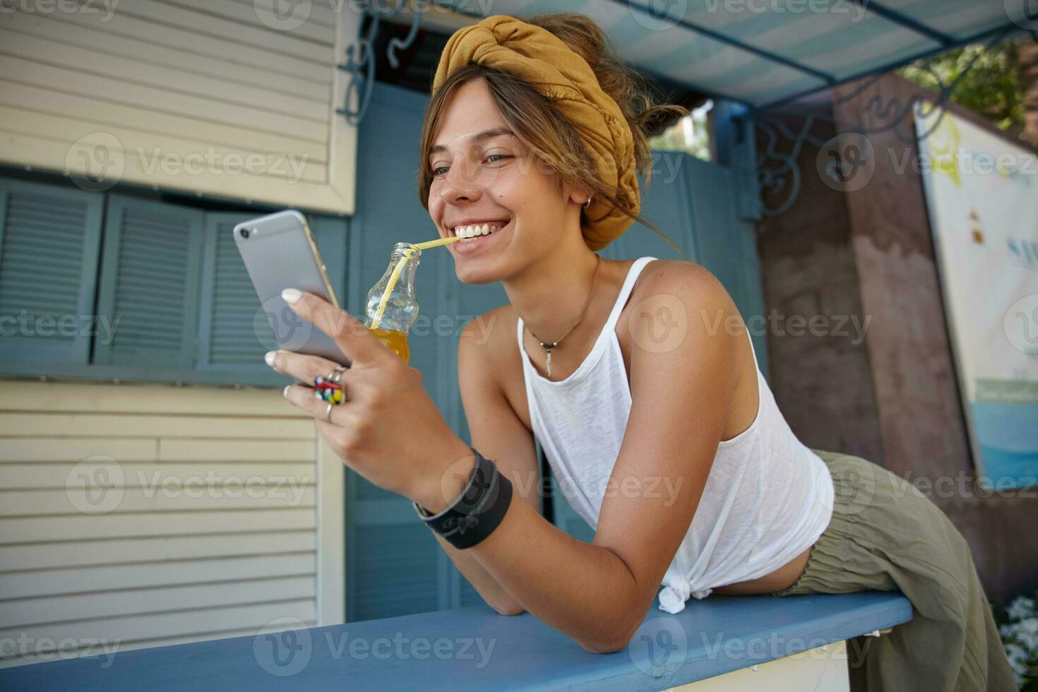Portrait of attractive young brunette female wearing white top and olive skirt posing over summer terrace, drinking lemonade with straw and typing message to her friend photo