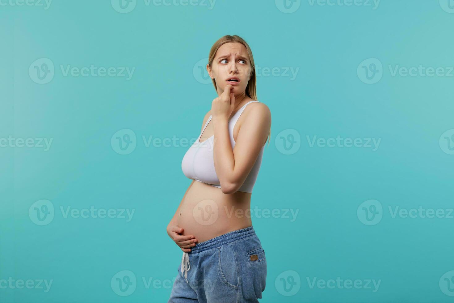 Frightened young blonde long haired woman being pregnant while standing over blue background, frowning eyebrows while looking scaredly aside and hugging her belly photo