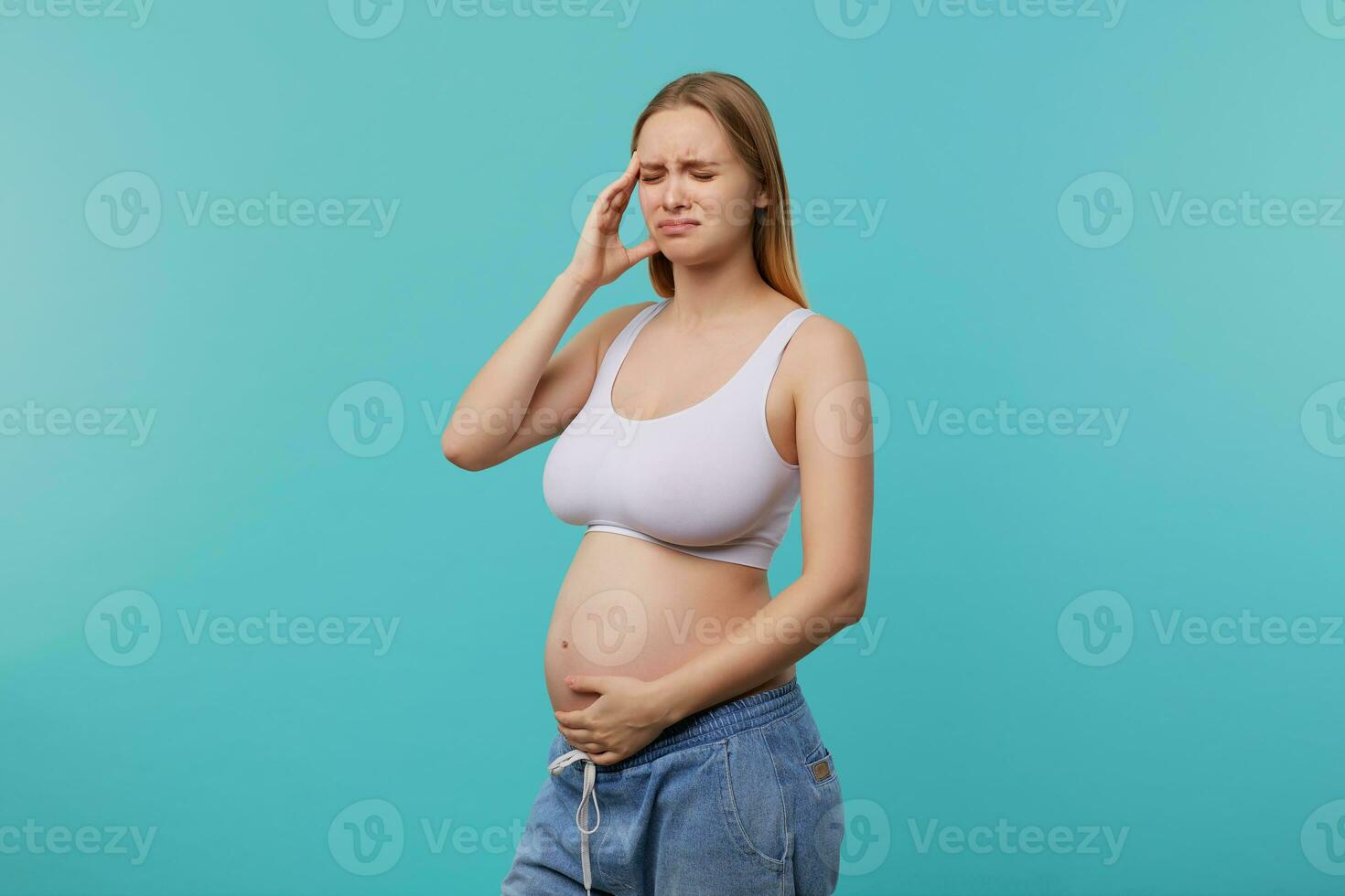 Stressed young blonde long haired future mom keeping raised hand on her belly and frowning sadly face with closed eyes, standing against blue background photo