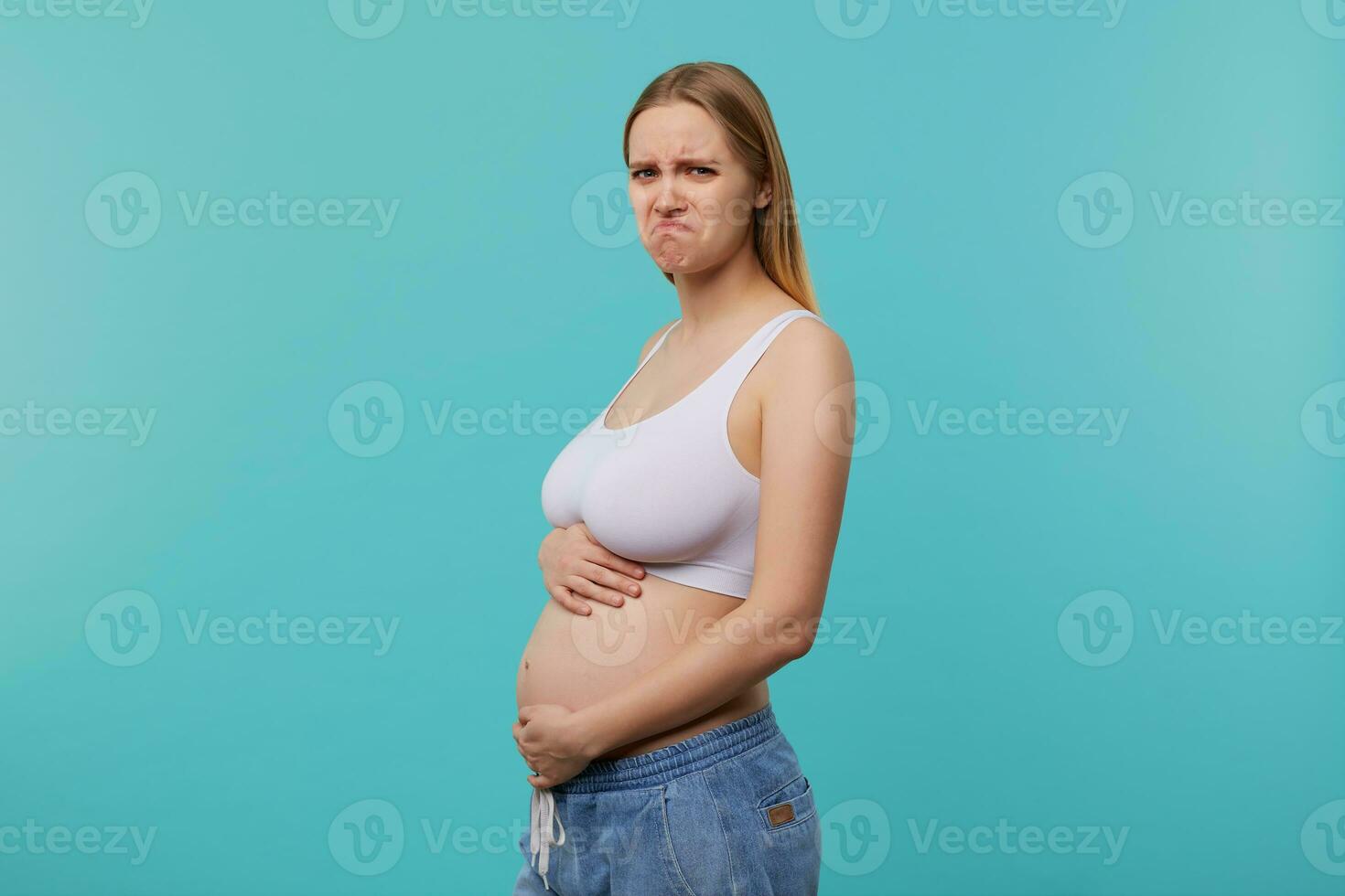 Indoor photo of young white-headed woman with casual hairstyle being pregnant while posing over blue background, twisting her mouth while looking sadly at camera