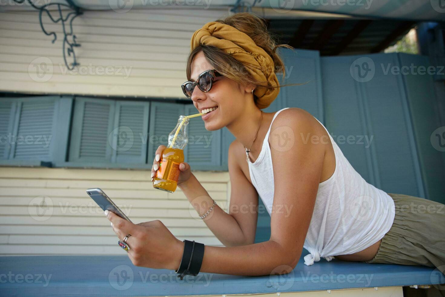 Outdoor shot of pretty young brunette lady in trendy sunglasses leaning on wooden stand, wearing casual clothes and mustard headband, drinking lemonade and checking social networks on smartphone photo