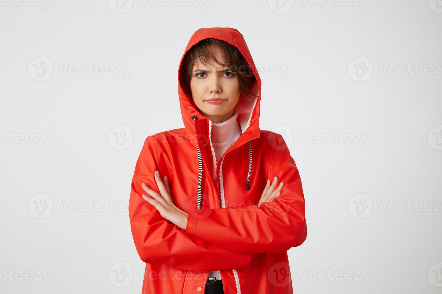 Portrait of young unhappy with rainy weather nice short haired lady dressed in red rain coat, with a hood on his head, looking away with sad expression, standing over white background. photo