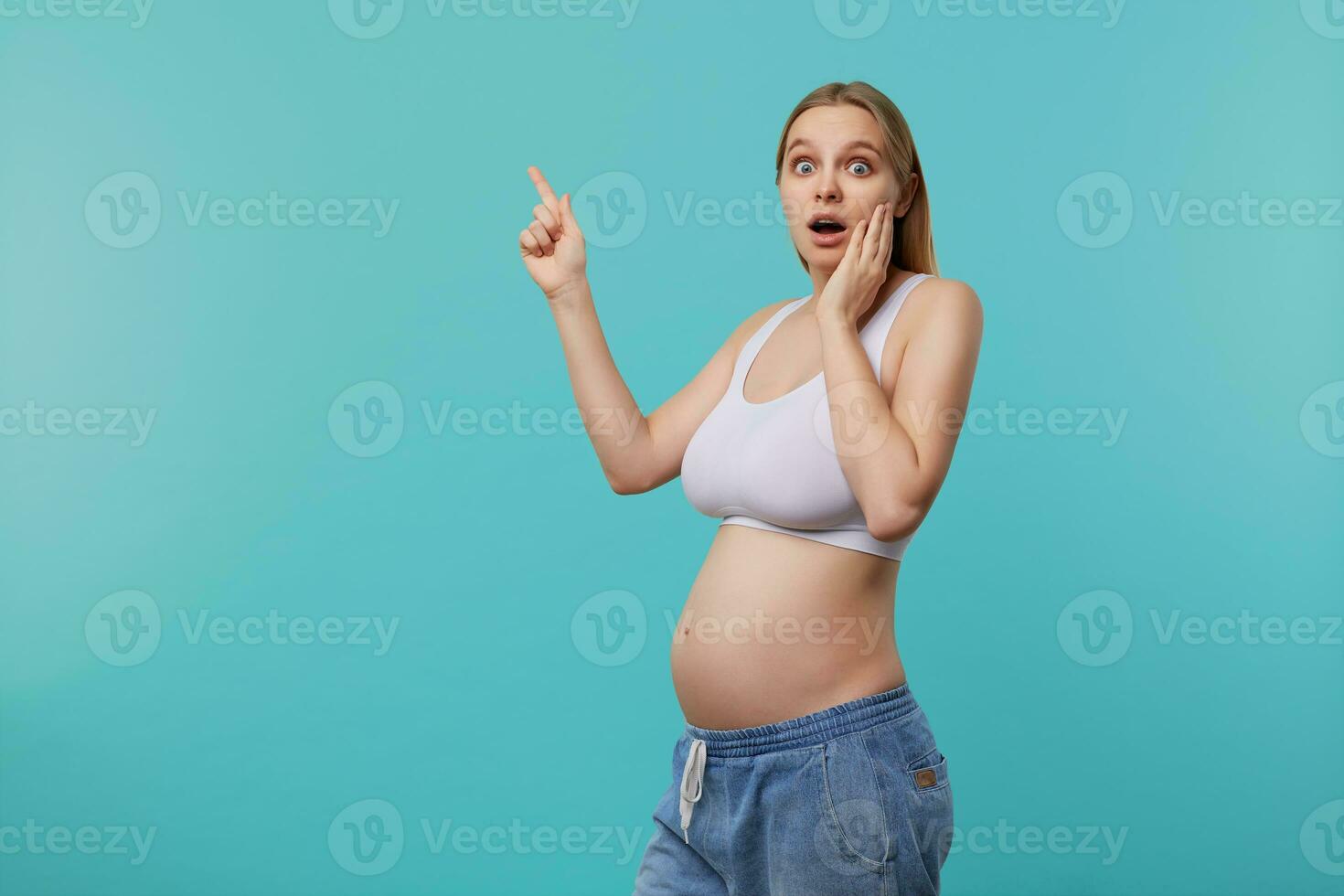 Shocked young blue-eyed blonde long haired female holding raised palm on her cheek while looking amazedly at camera, being pregnant while posing over blue background photo